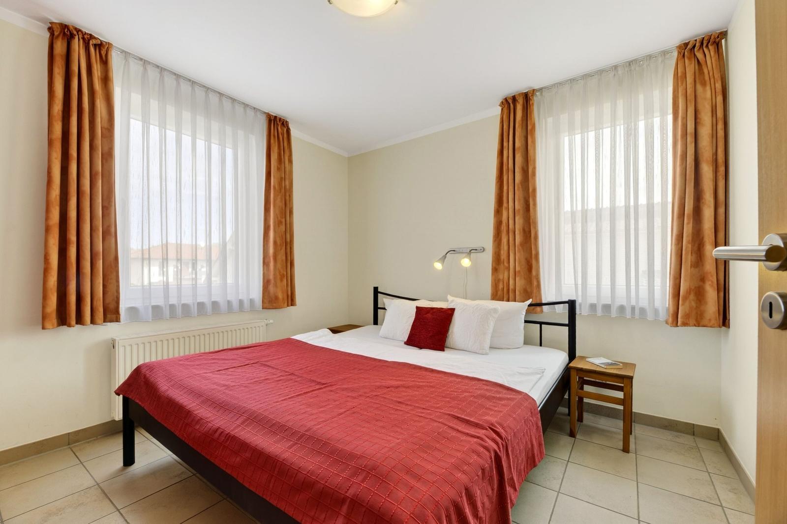 Schlafzimmer mit Doppelbett, zwei Fenstern und Holztisch