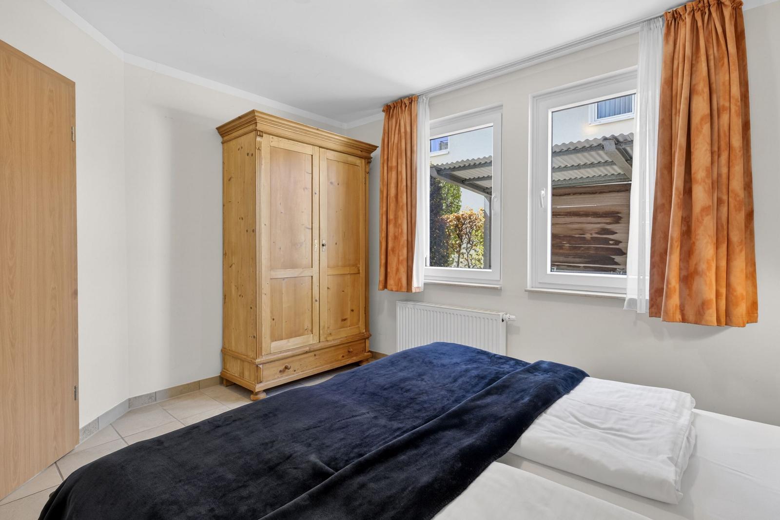 Schlafzimmer mit Bett, Holzschrank und Fenstern mit Vorhängen.