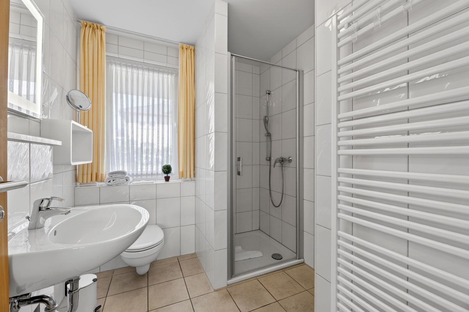 Badezimmer mit Dusche, Waschbecken und Toilette. Fenster mit gelben Vorhängen.