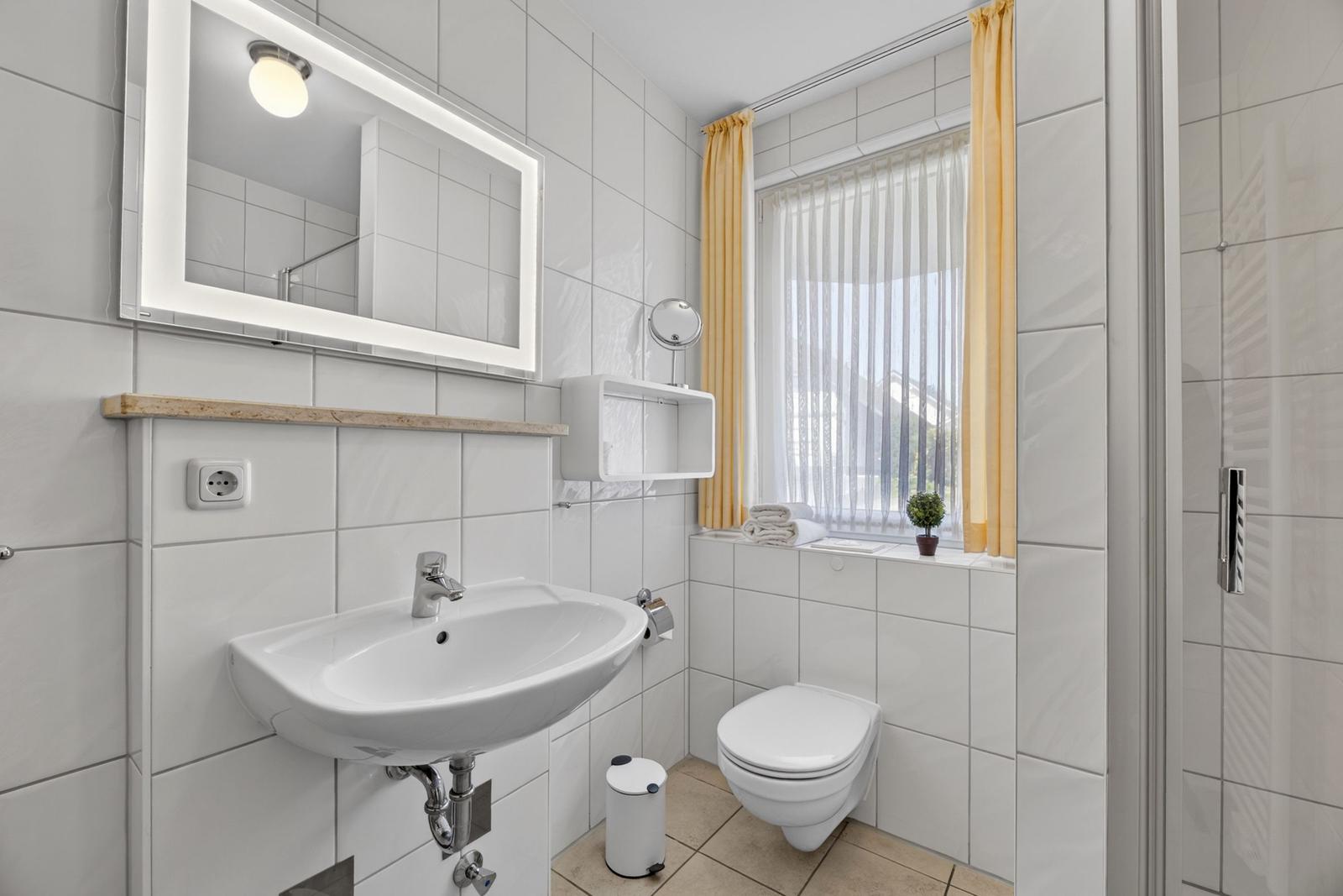 Badezimmer mit Waschbecken, Spiegel, WC und Dusche. Fenster mit gelben Vorhängen.