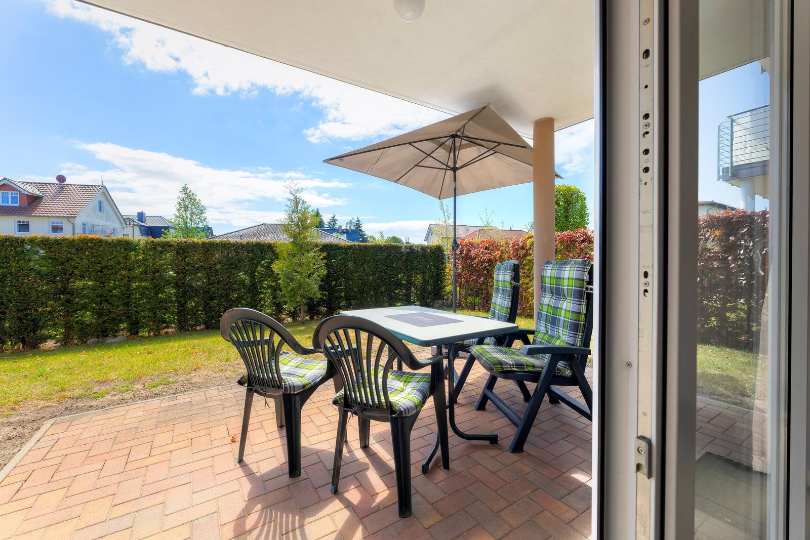 Terrasse mit Tisch, vier Stühlen und Sonnenschutz. Hintergrund: grüner Heckenzaun und Häuser.