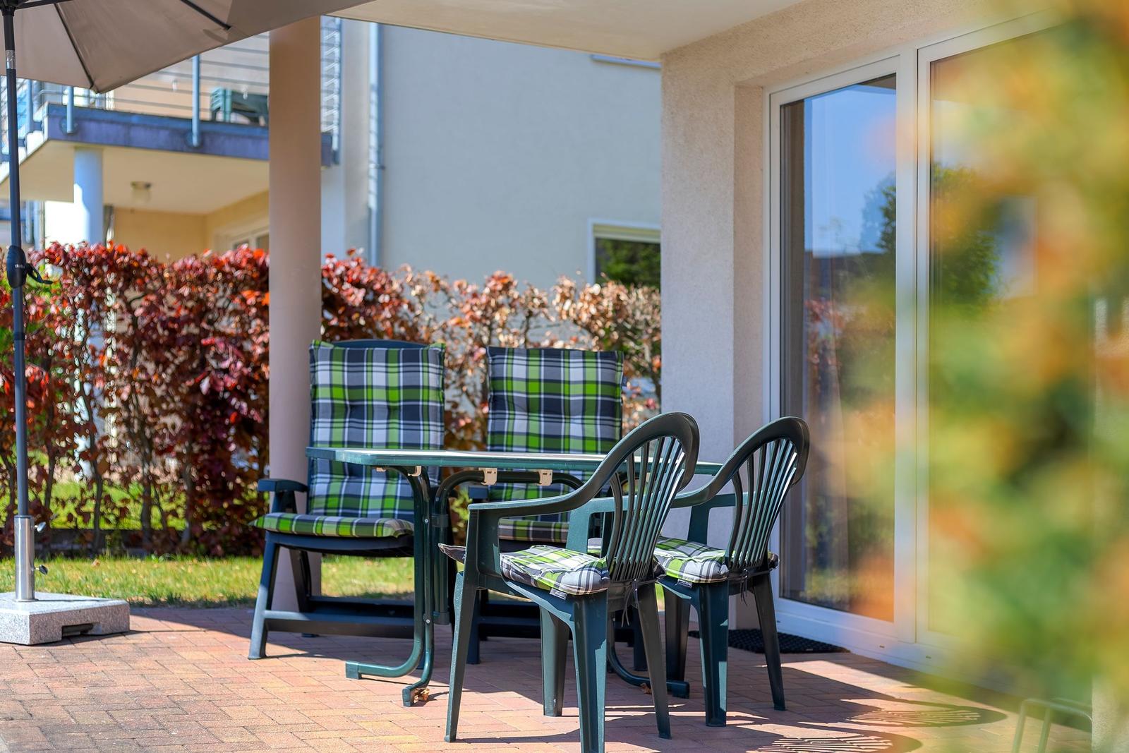 Terrasse mit Tisch, vier Stühlen und Sonnenschutz. Hintergrund: Beete und Glasfront.