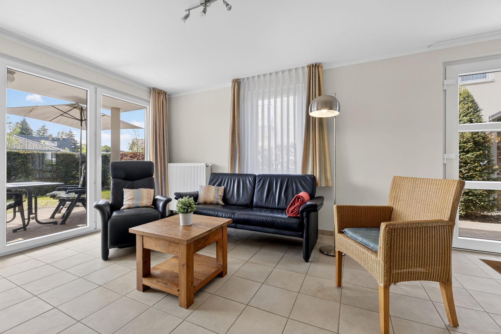 Wohnzimmer mit Sofa, Sessel und Tisch. Durchgänge zu Terrasse und Garten.