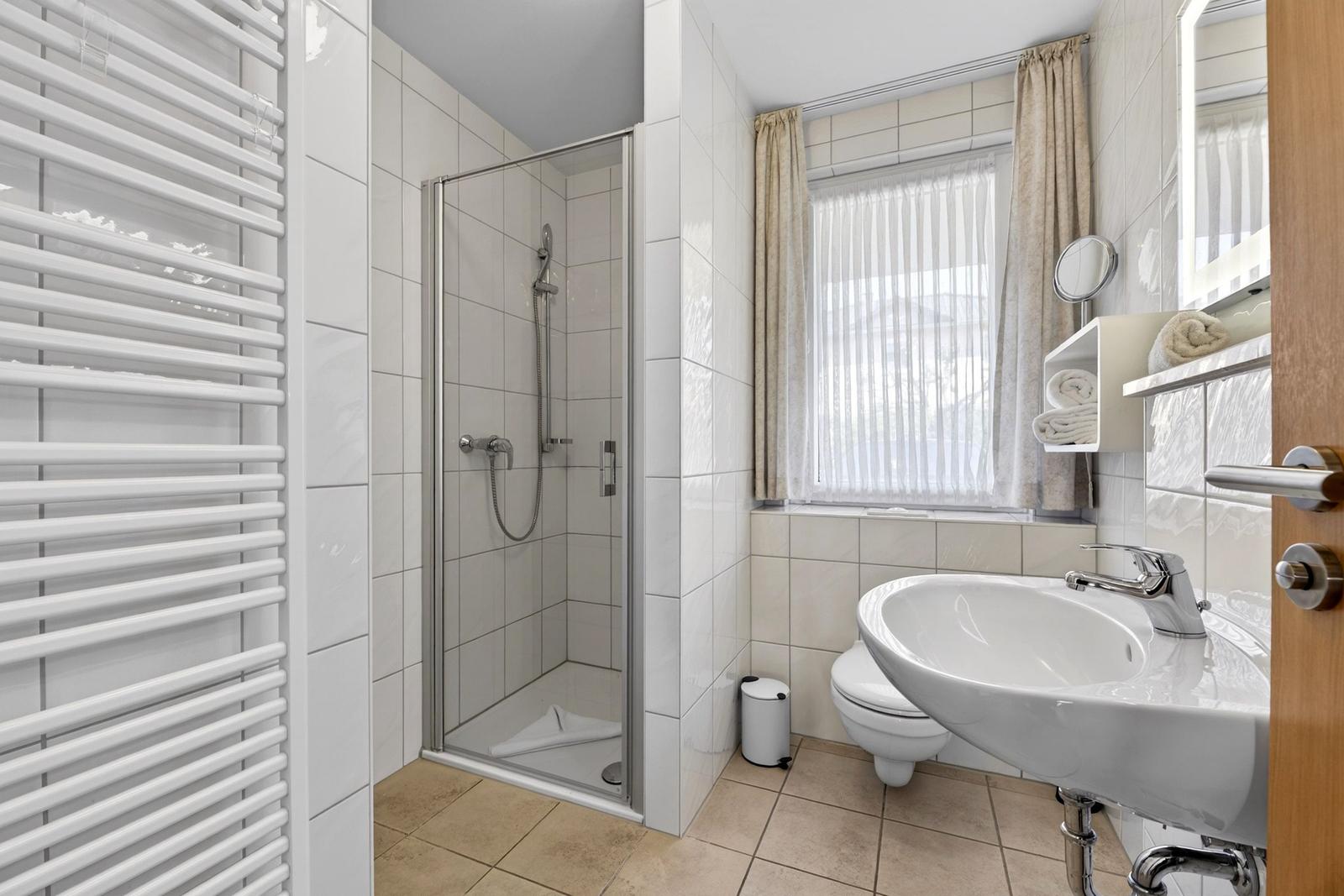 Badezimmer mit Dusche, Waschbecken und Toilette. Fenster mit Vorhängen.