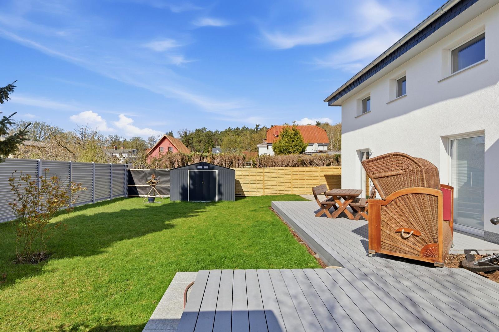 Hinterhof mit Terrasse, Garten und Holzstühlen. Weiße Hauswand und Holzgitter.