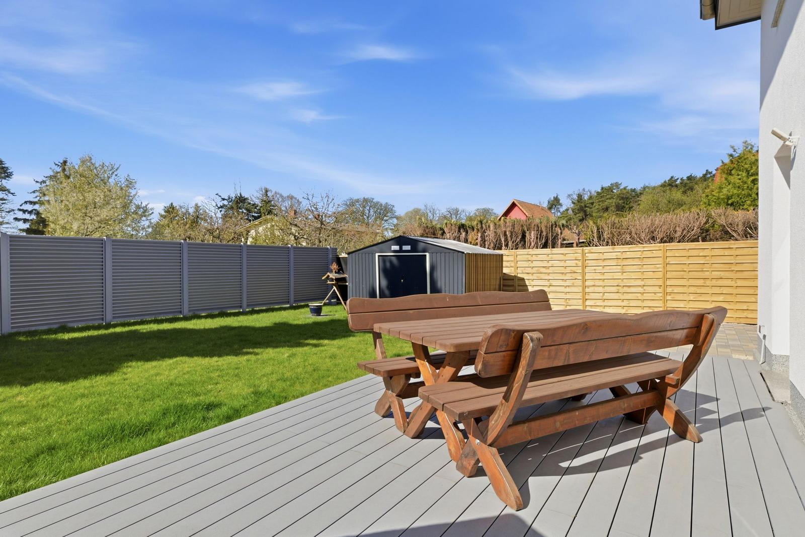 Holztisch und Bänke auf Terrasse mit Garten und Zaun im Hintergrund.