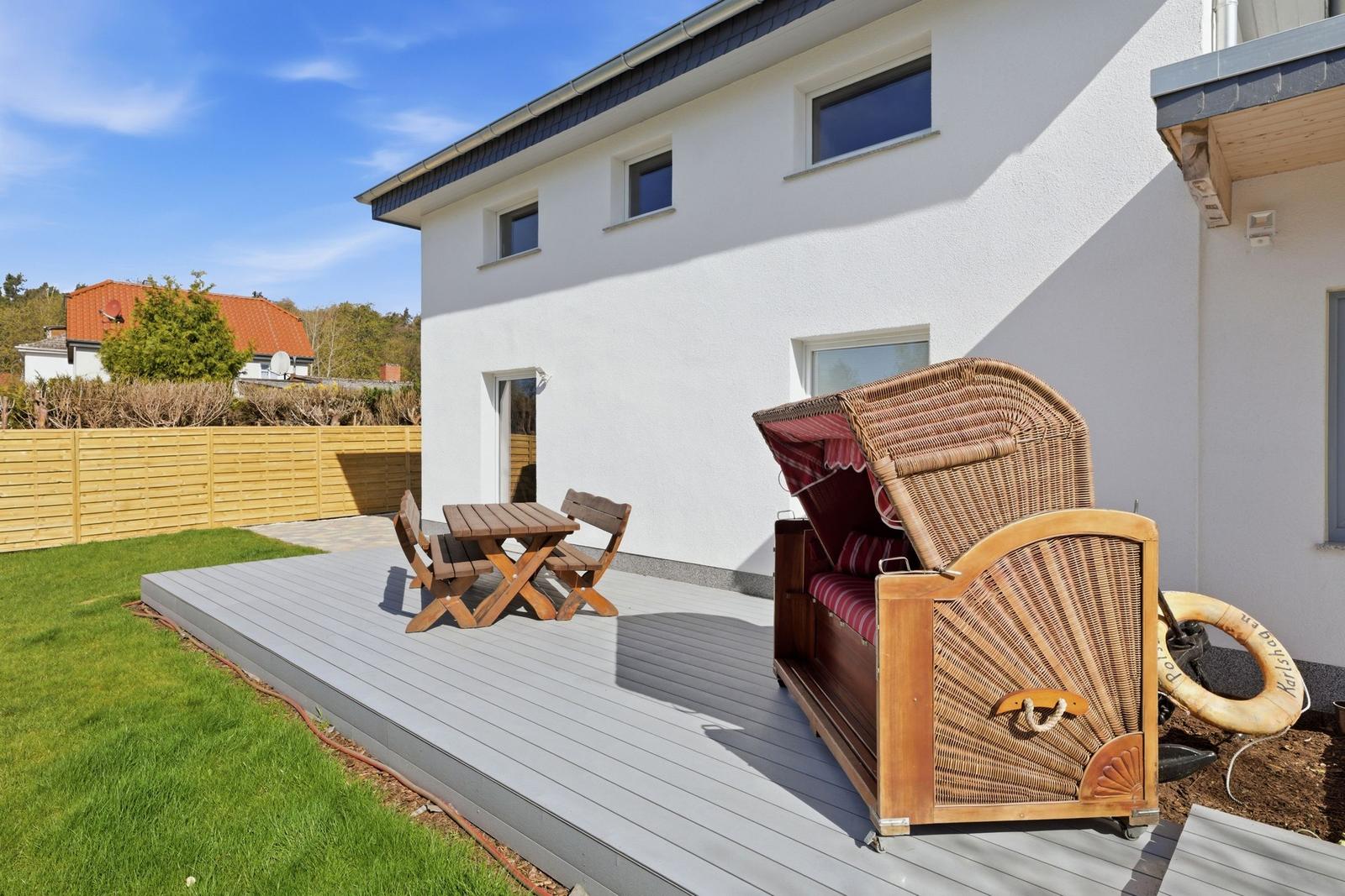 Terrasse mit Holztisch, Bistrostühlen und Strandkorb. Hintergrund: weißes Haus, Holzzaun und Gras.