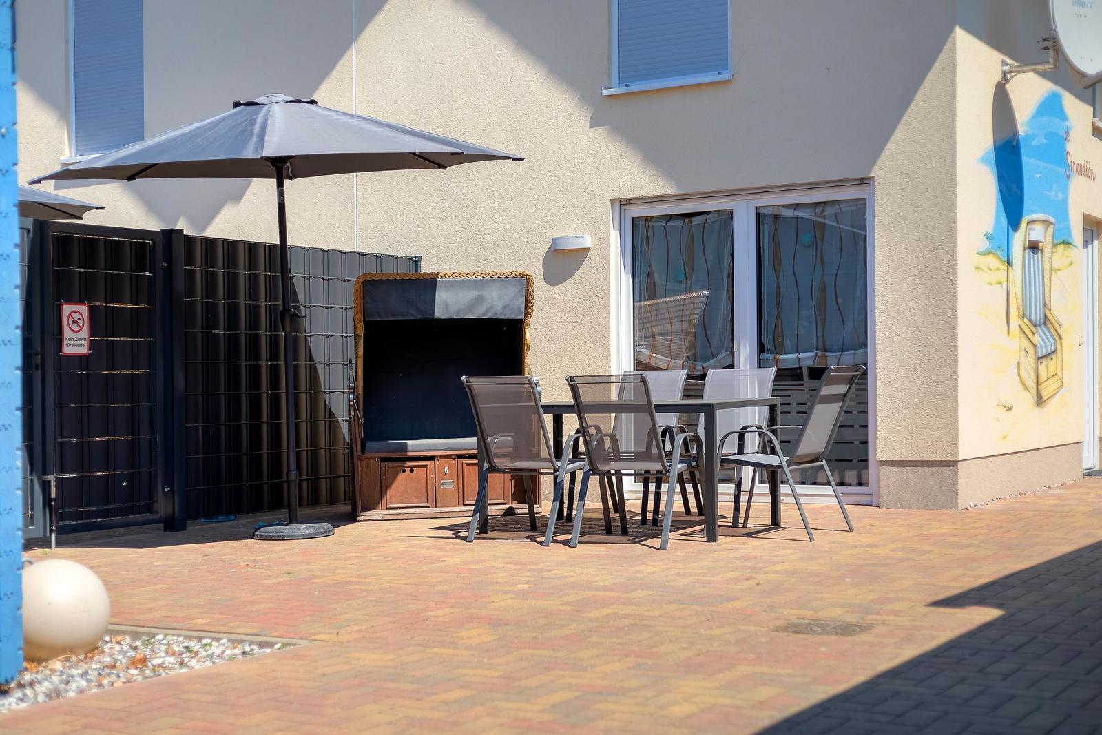 Terrasse mit Tisch, Stühlen und Sonnenschirm. Hintergrund: Gebäude mit Fenster und Wandmalerei.
