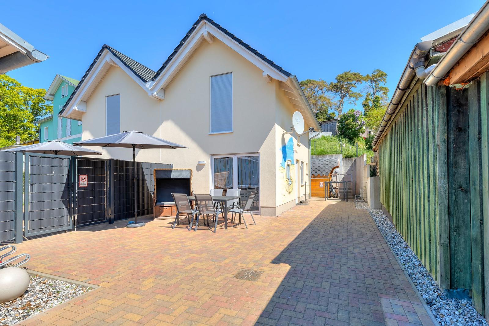 Haus mit Terrasse, Tisch, Stühlen und Sonnenschirmen. Hinterhof mit Holzzaun und Garten.