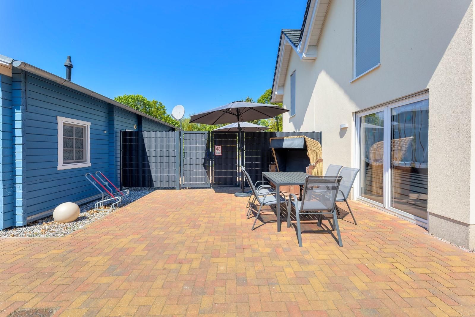 Terrasse mit Tisch, Stühlen und Sonnenschirm neben Haus und blauem Anbau.