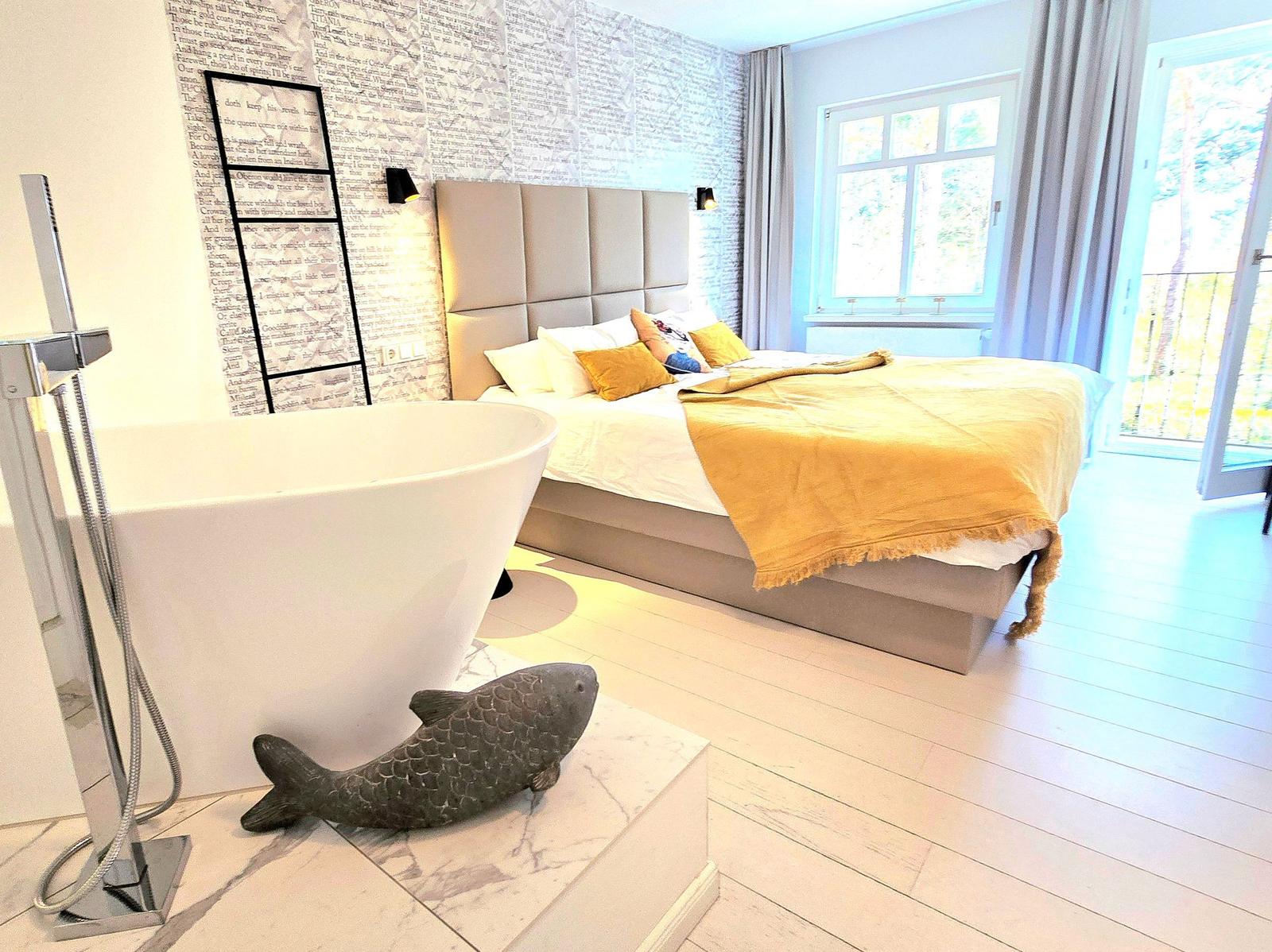 Schlafzimmer mit freistehender Badewanne und Holzboden. Wand mit Buchstabenmuster und Fenster mit Blick.