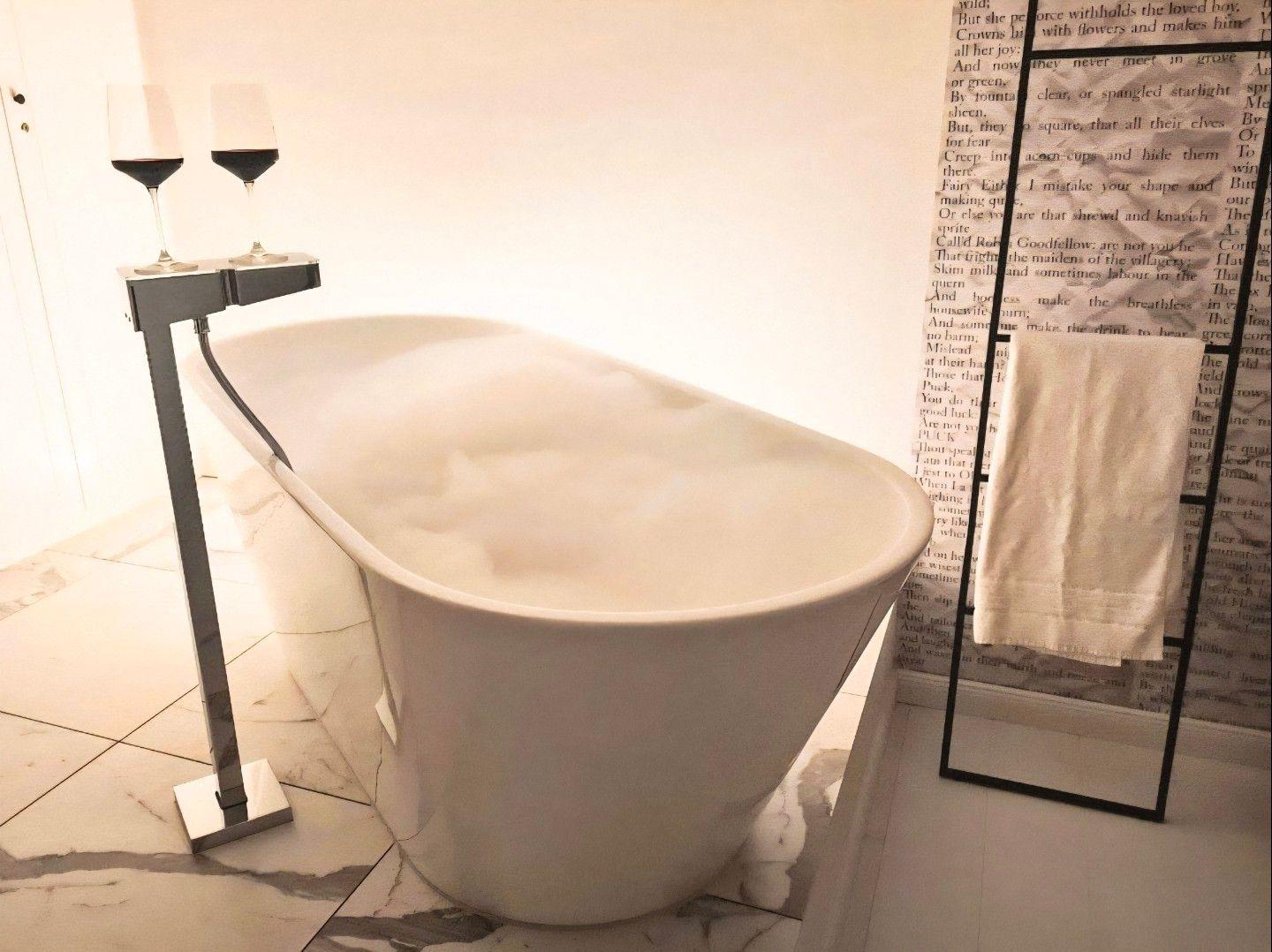 Freistehende Badewanne mit Schaum, zwei Weingläser und Handtuchhalter.