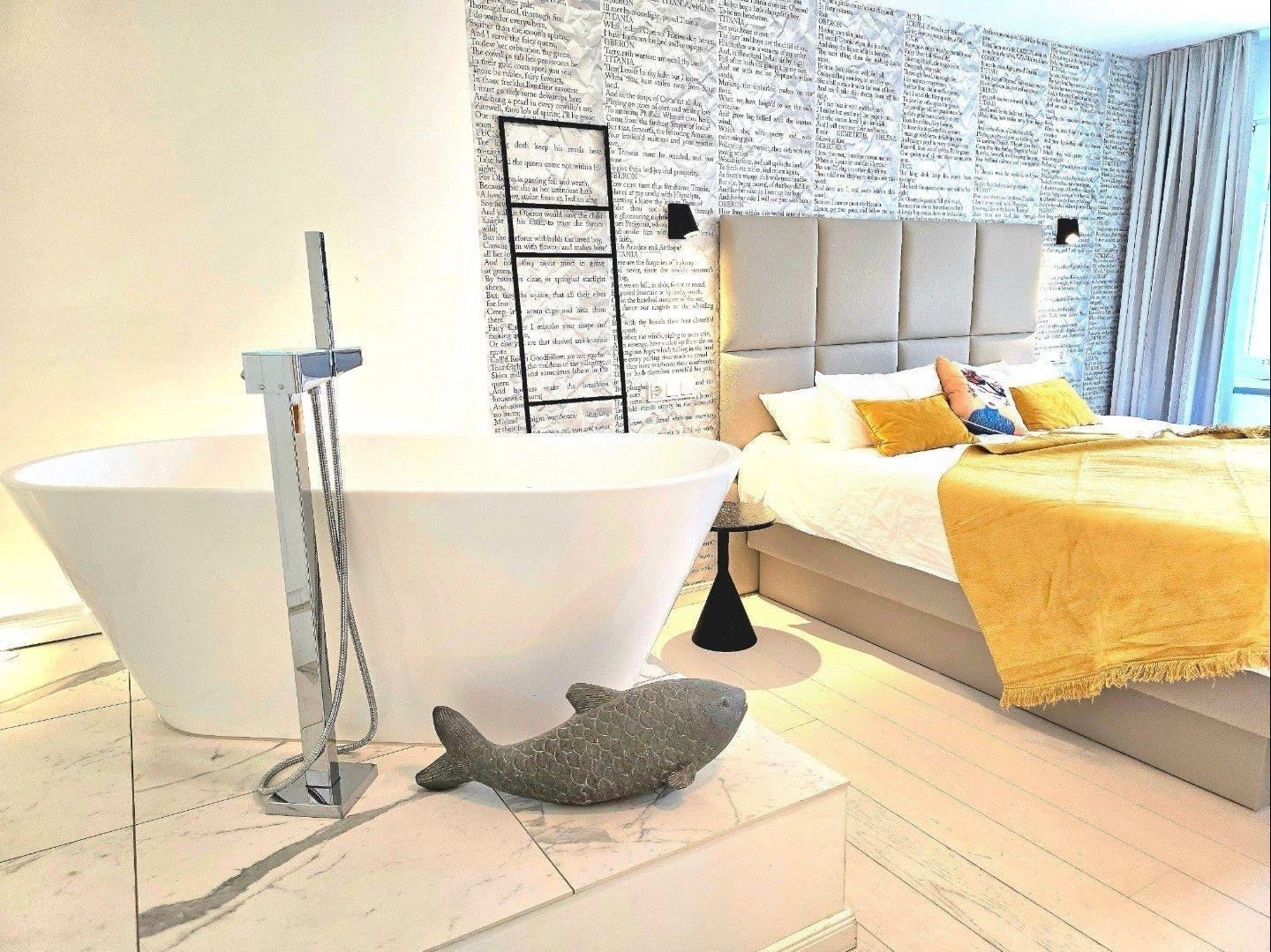 Schlafzimmer mit Badewanne, Textilmusterwand und gelbem Bettbezug.