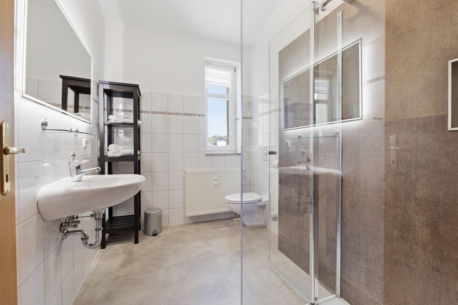 Badezimmer mit Dusche, Waschbecken und Toilette. Fenster und Heizung sichtbar.