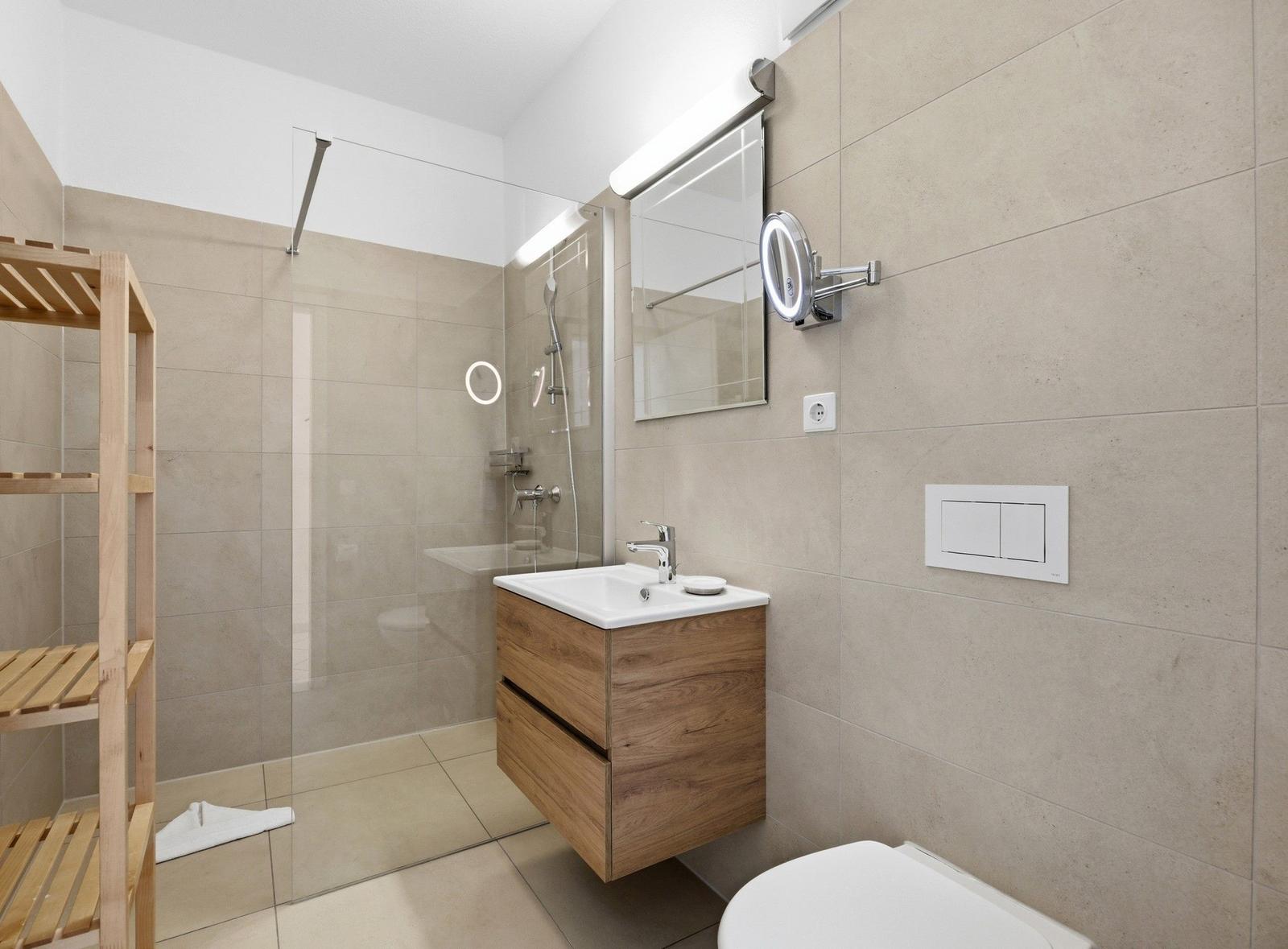 Bathroom with shower, sink, and toilet. Wooden shelf and mirror with lighting.