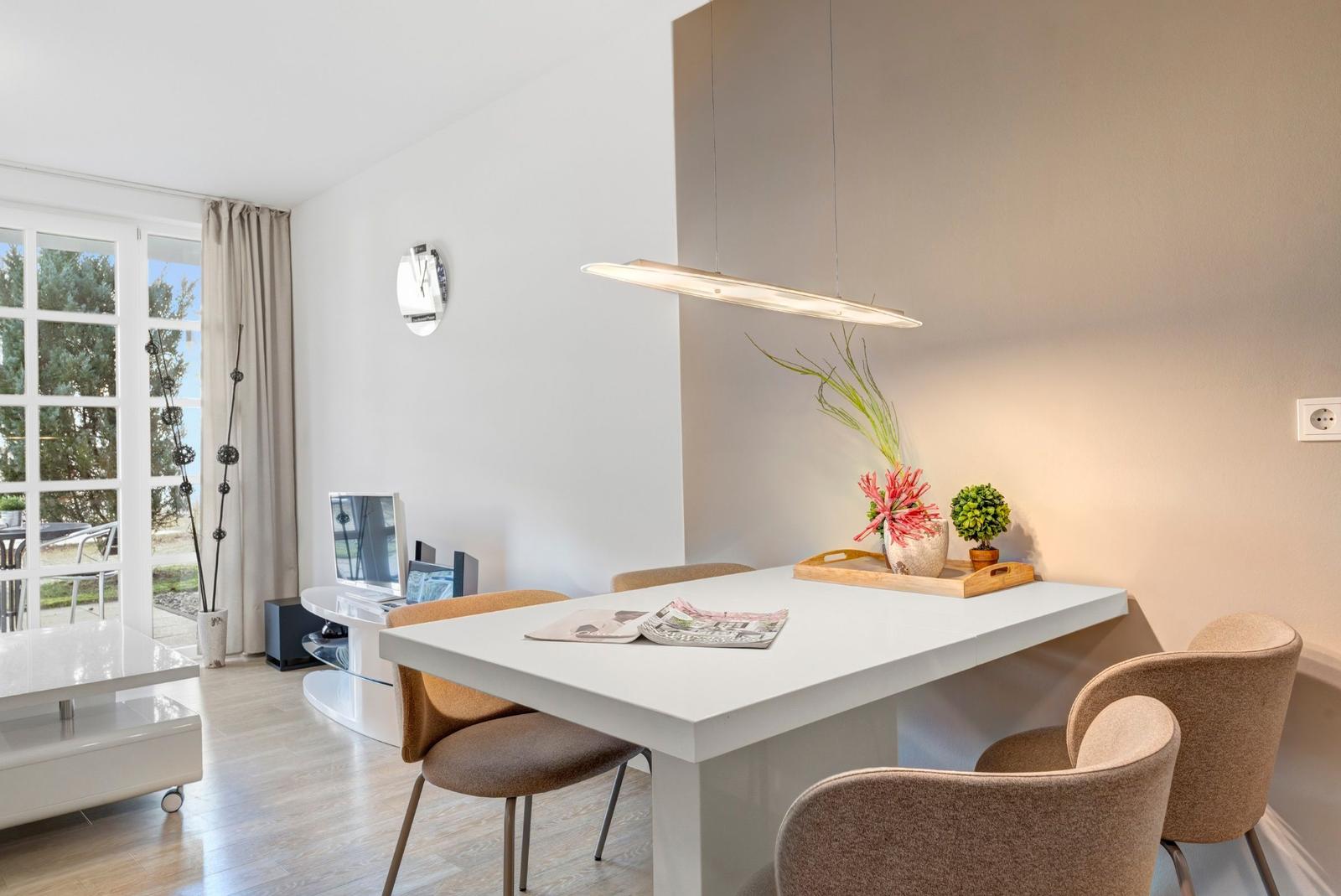 Essbereich mit weißem Tisch und beigen Stühlen, daneben Wohnbereich mit TV und Fenster.
