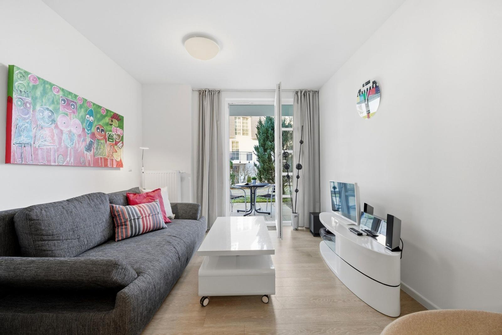 Wohnzimmer mit Sofa, Couchtisch und TV-Möbel. Durchgang zur Terrasse mit Tisch und Stühlen.