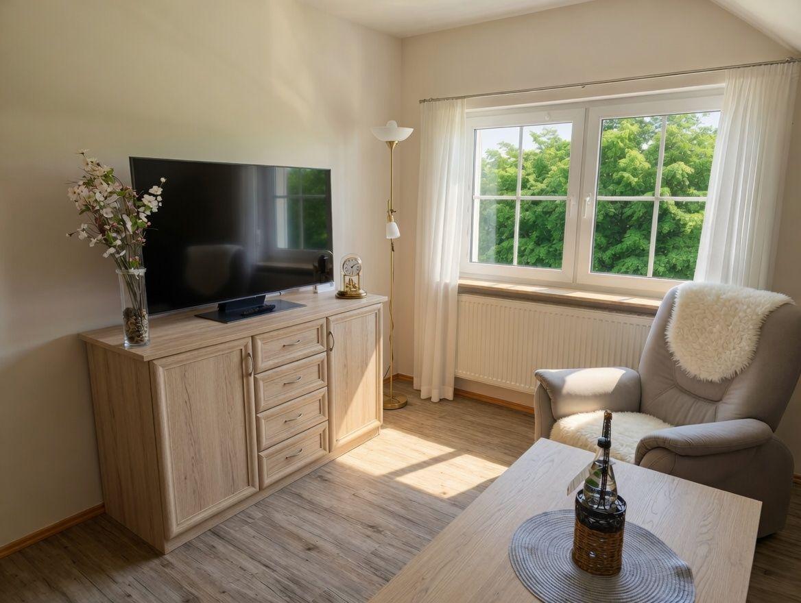 Ein Wohnzimmer mit Fernseher, Sessel und Tisch. Durch ein Fenster blickt man auf Bäume.