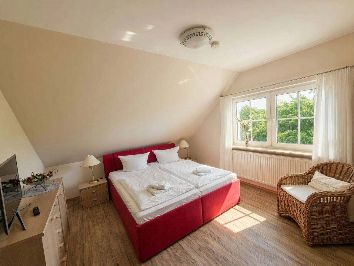 Schlafzimmer mit rotem Bett, Wicker-Sessel und Fenster mit Blick auf Grün.