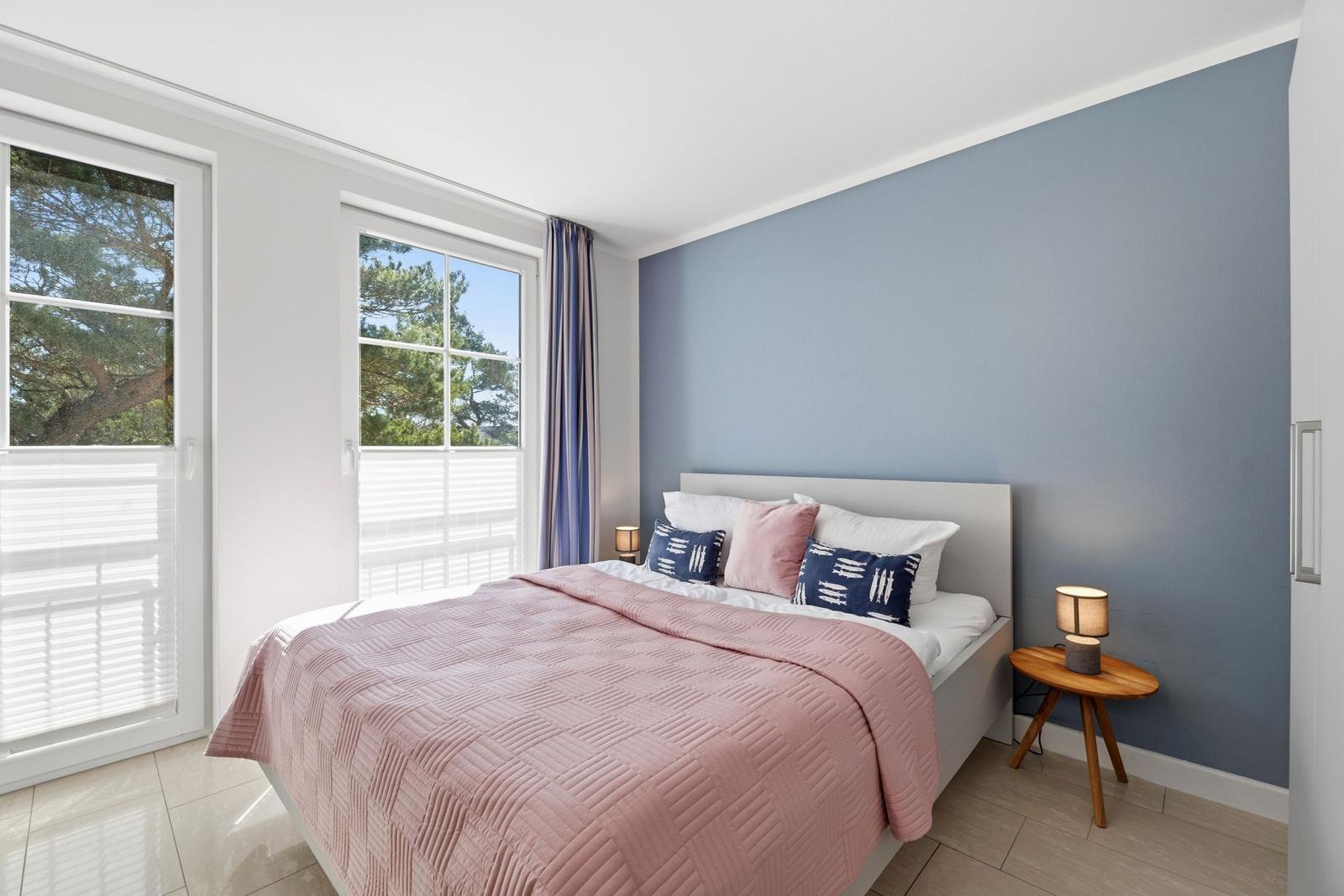 Schlafzimmer mit Bett, blauer Wand und Fenstern zum Garten.