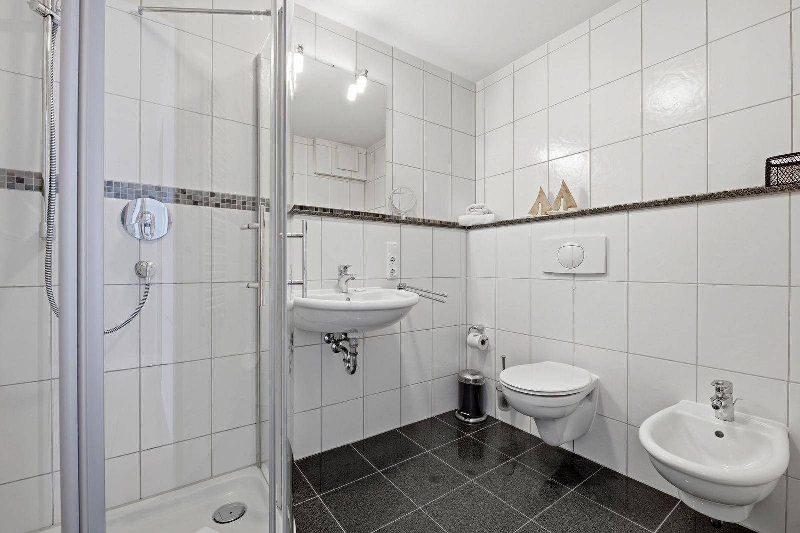 Badezimmer mit Dusche, Waschbecken, Toilette und Bidet. Weiße Fliesen und dunkler Boden.
