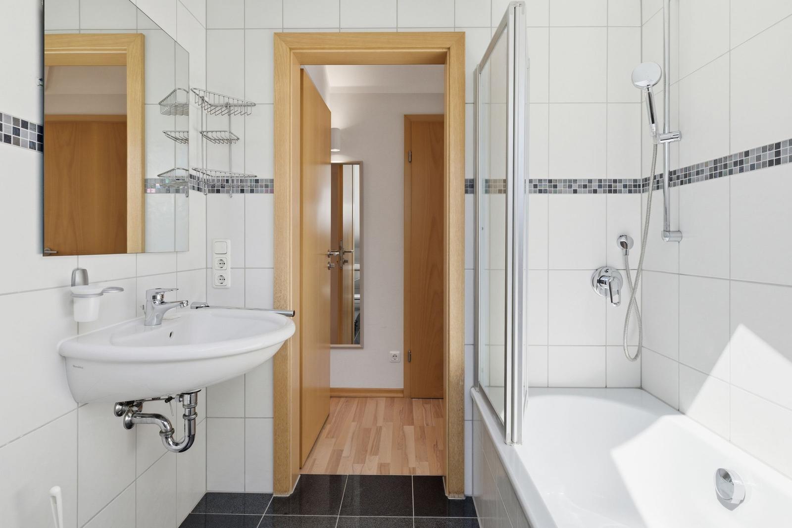 Badezimmer mit Dusche, Waschbecken und Spiegel. Holzfußboden im angrenzenden Raum.