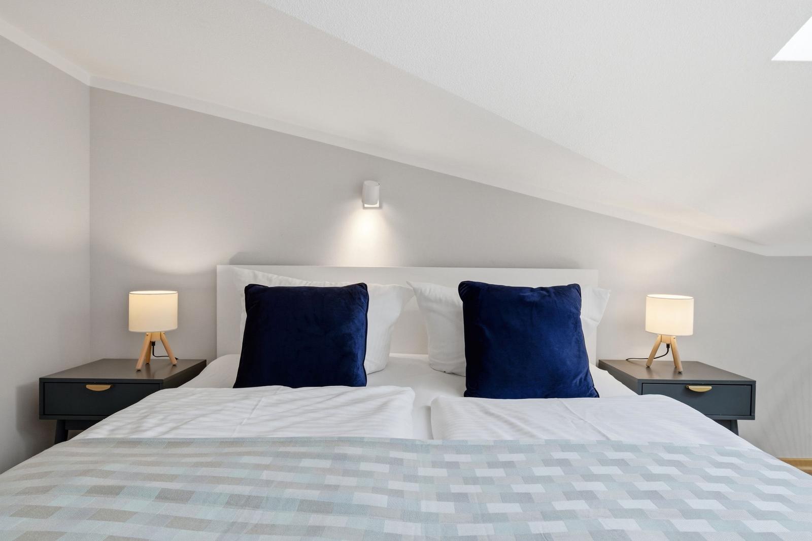 Schlafzimmer mit Bett, zwei Nachttischen und blauen Kissen unter schrägem Dach.