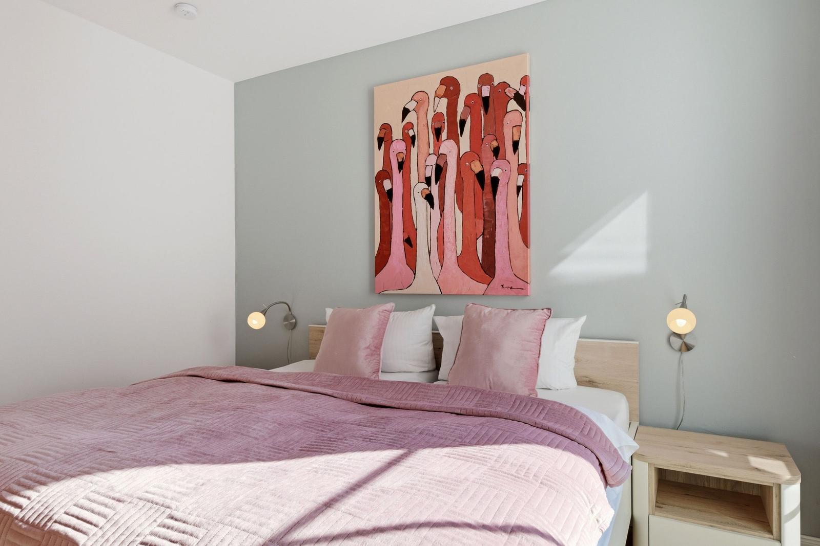 Schlafzimmer mit Bett, rosa Bettwäsche, Wandbild mit Flamingos und Nachttischen.