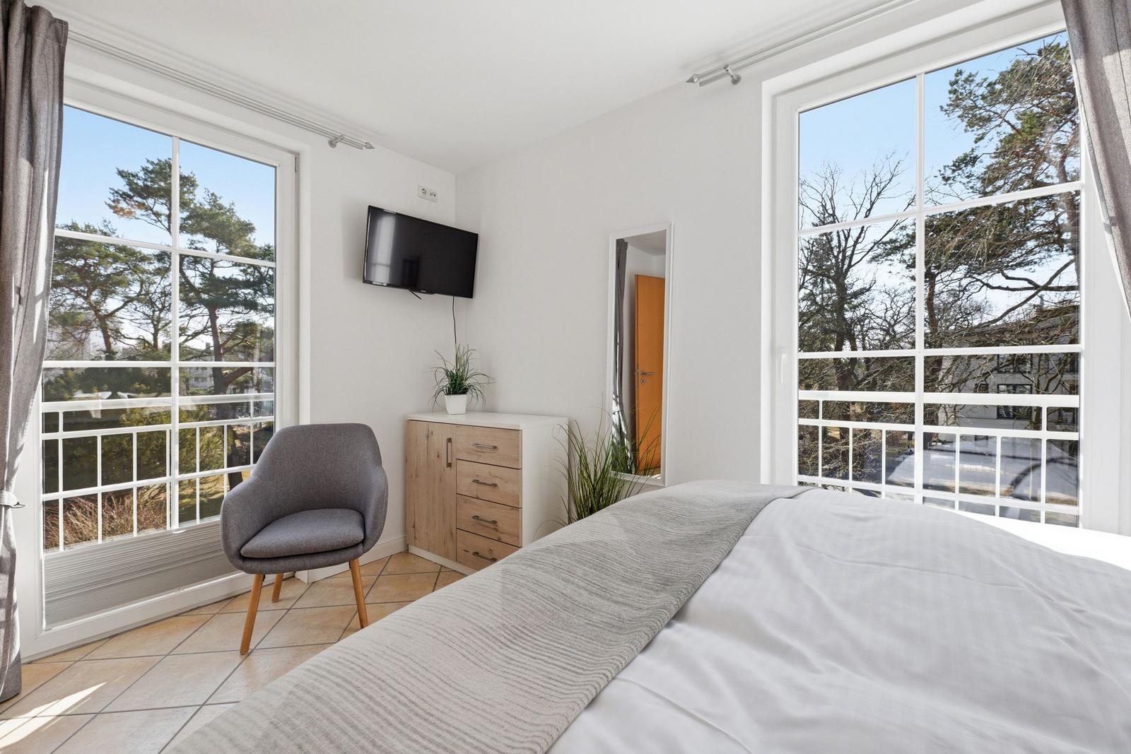 Schlafzimmer mit Bett, Stuhl, Schrank und TV. Große Fenster mit Blick auf Bäume.