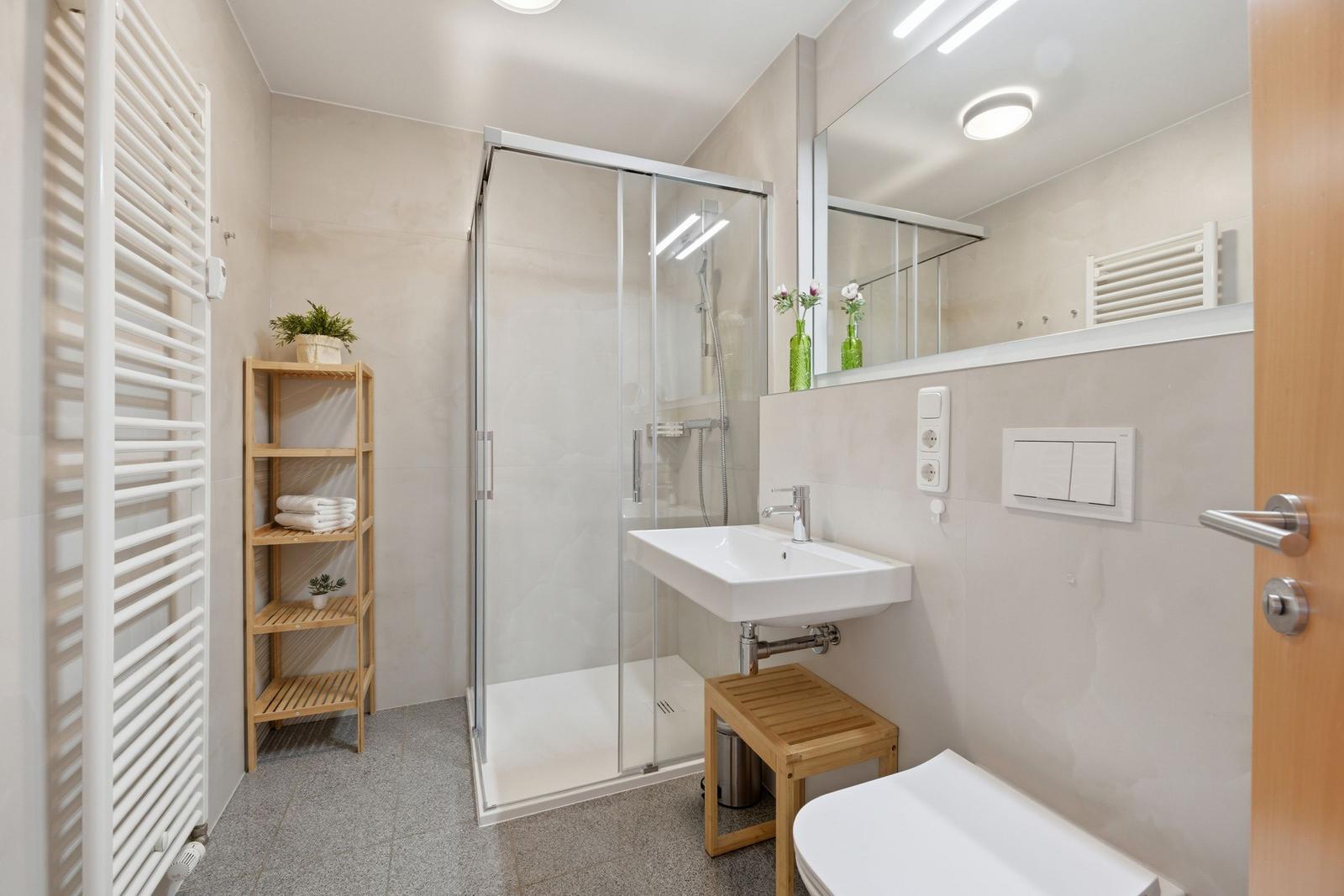 Badezimmer mit Dusche, Waschbecken und Toilette. Holzregal und Heizkörper sichtbar.