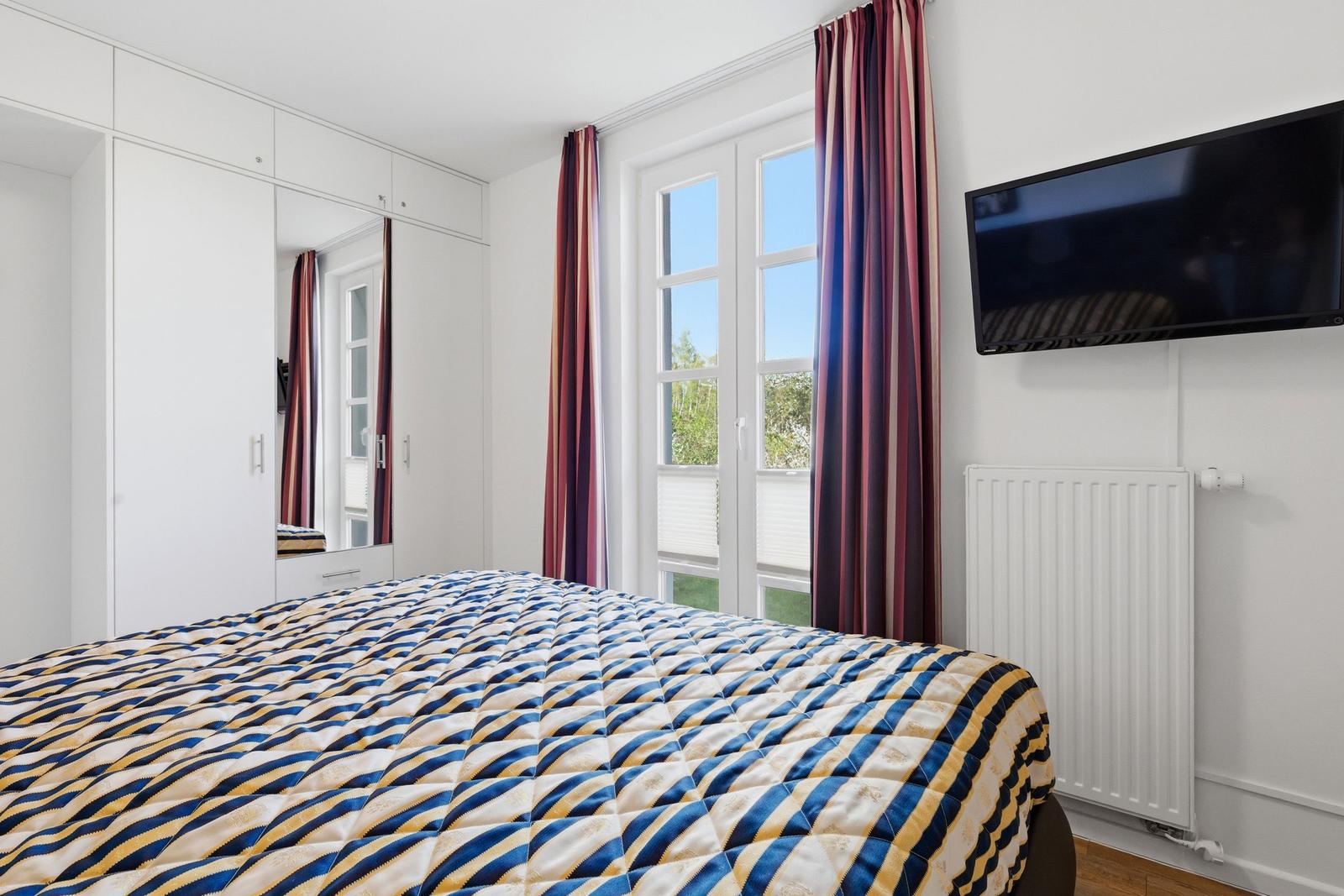 Schlafzimmer mit Bett, Fenster, TV und Kleiderschrank.
