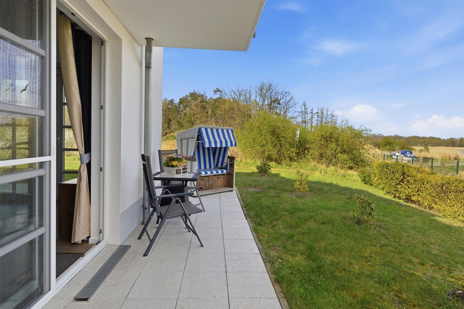 Terrasse mit Strandkorb und Tisch, Blick auf Grünland und Wasser.