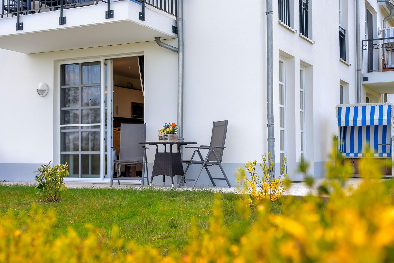 Außenbereich mit Tisch und Stühlen vor einem Haus mit Balkon und blau-weißem Sonnenschutz.
