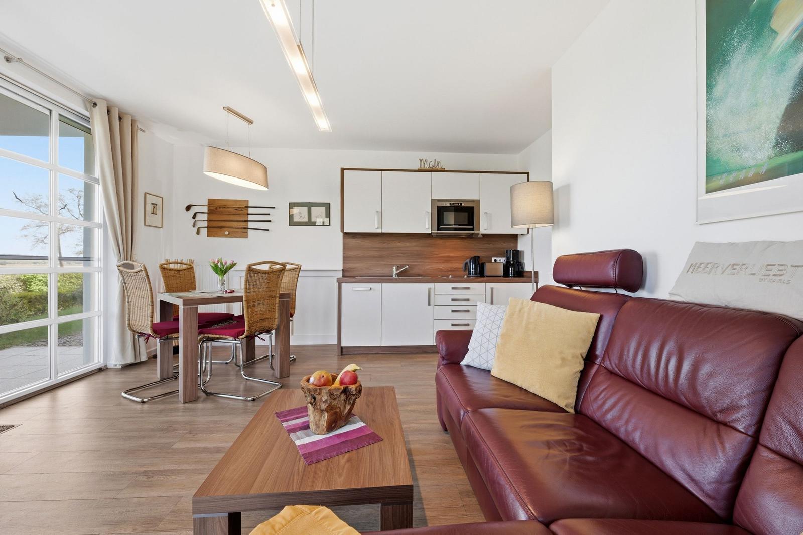 Wohnbereich mit Sofa, Esstisch und Küchenzeile. Große Fensterfront mit Blick nach draußen.