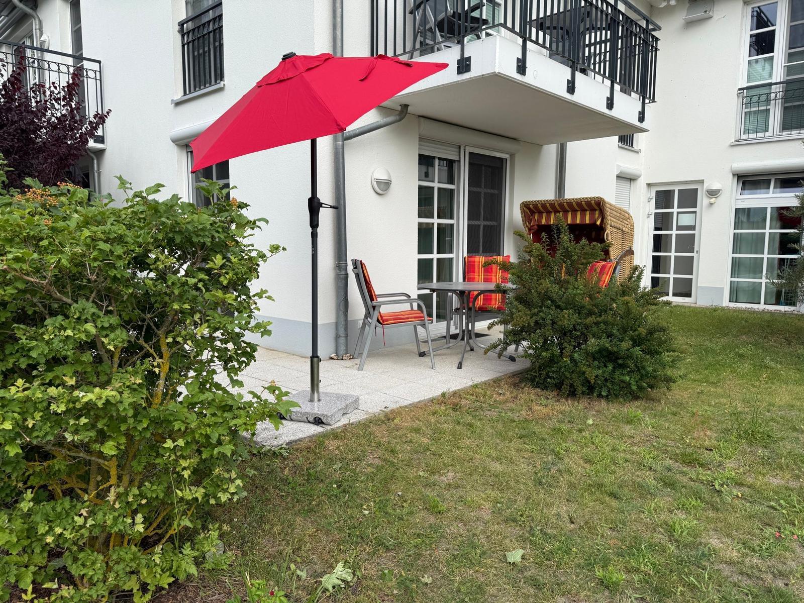 Terrasse mit rotem Sonnenschirm, Tisch und Stühlen. Grünflächen und Hecken umgeben den Außenbereich.