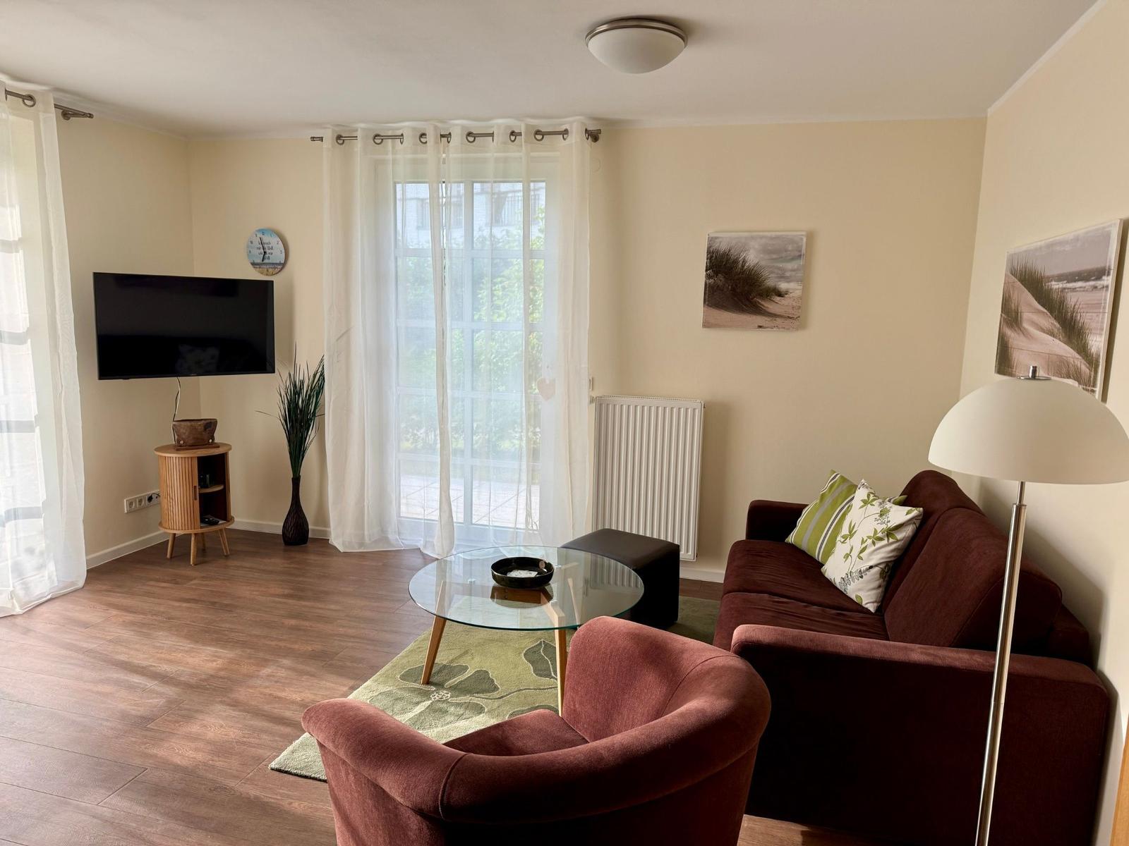 Wohnzimmer mit Sofa, Sessel, Glas-Tisch und Fernseher. Fenster mit weißen Vorhängen.
