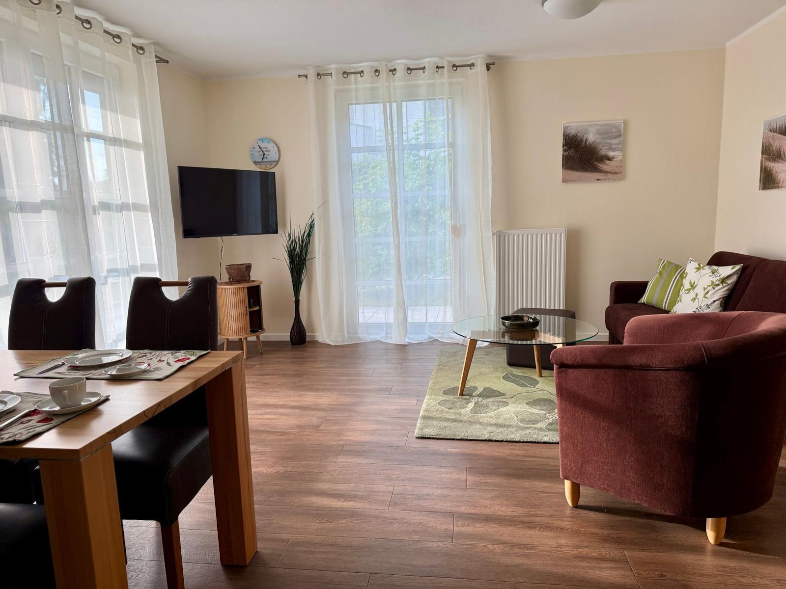 Wohn- und Essbereich mit Sofa, Tisch und TV. Holzboden und große Fenster.