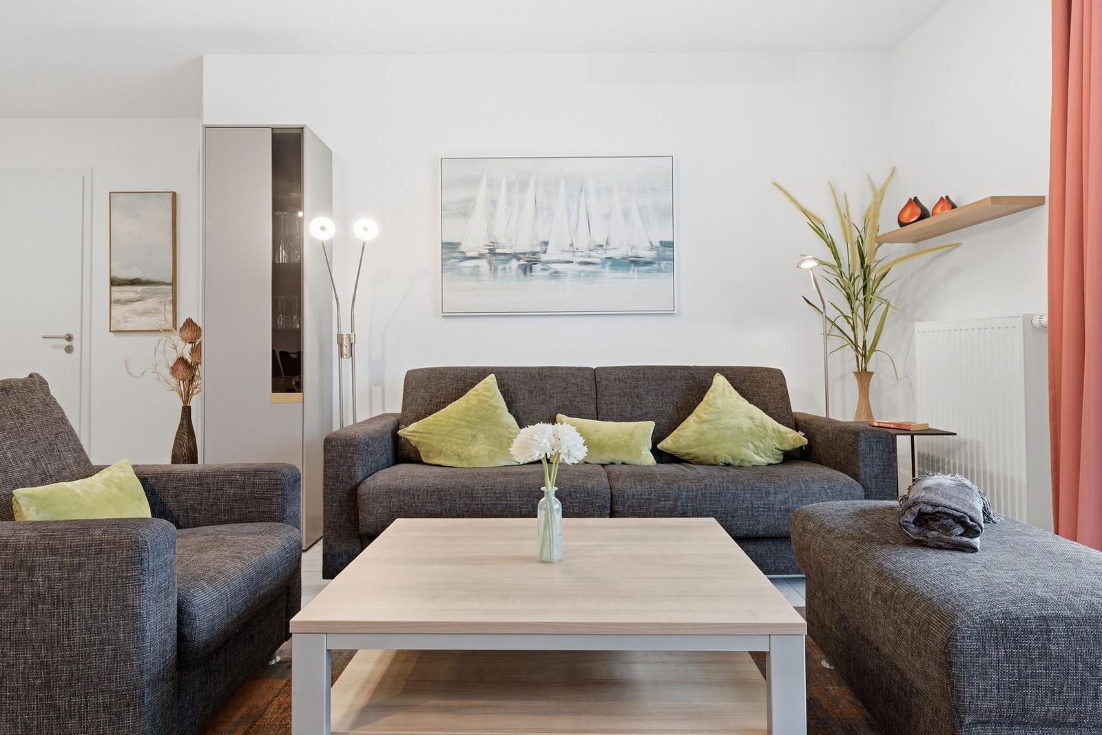 Wohnzimmer mit grauem Sofa, grünen Kissen und Holztisch. Wandbild mit Segelbooten.