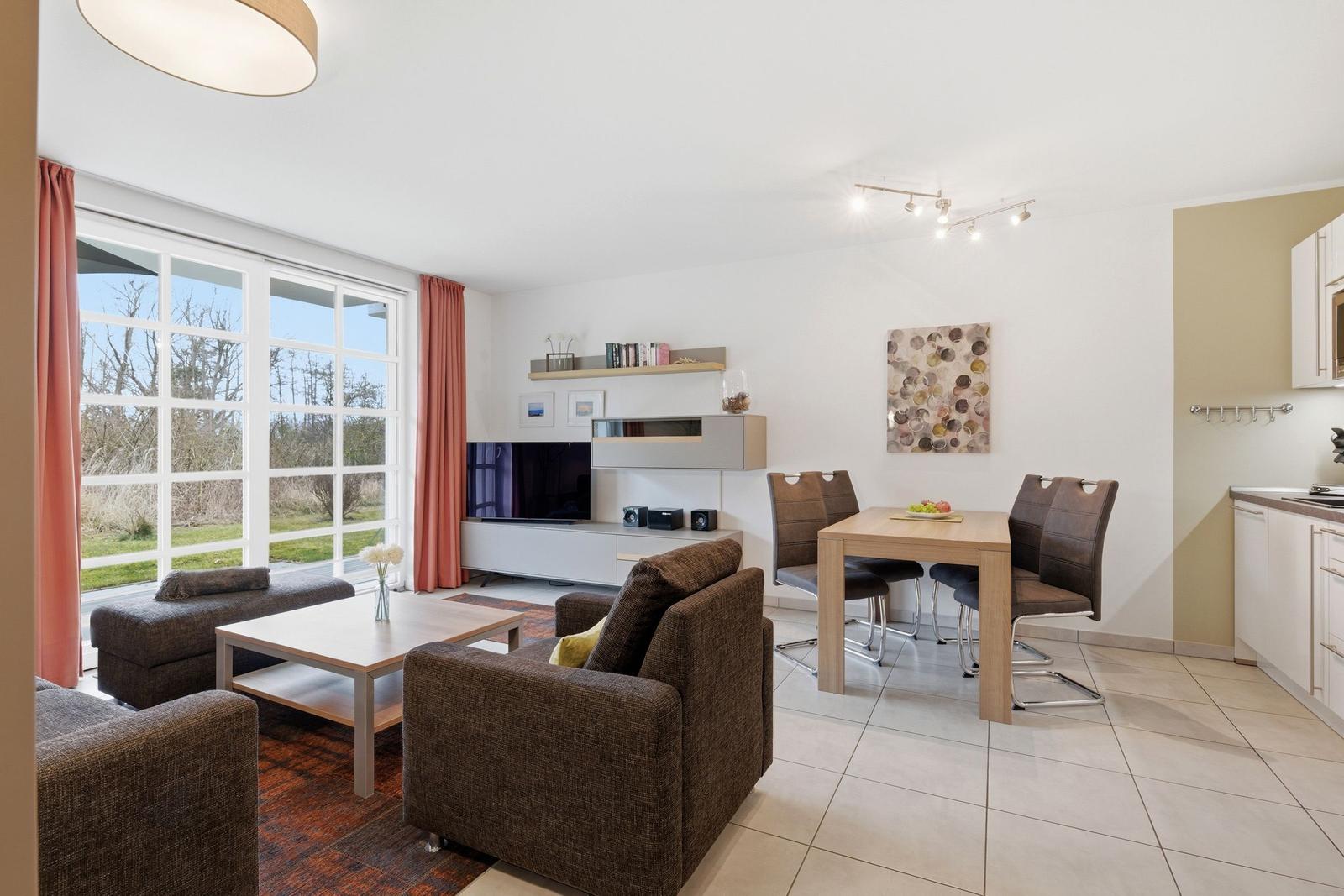 Wohn- und Essbereich mit Fernseher, Tisch und Stühlen, großen Fenstern und Küche im Hintergrund.