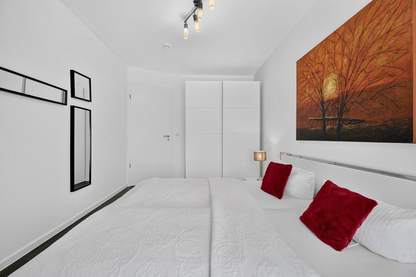 Schlafzimmer mit weißem Bett, roten Kissen und Wandbild.