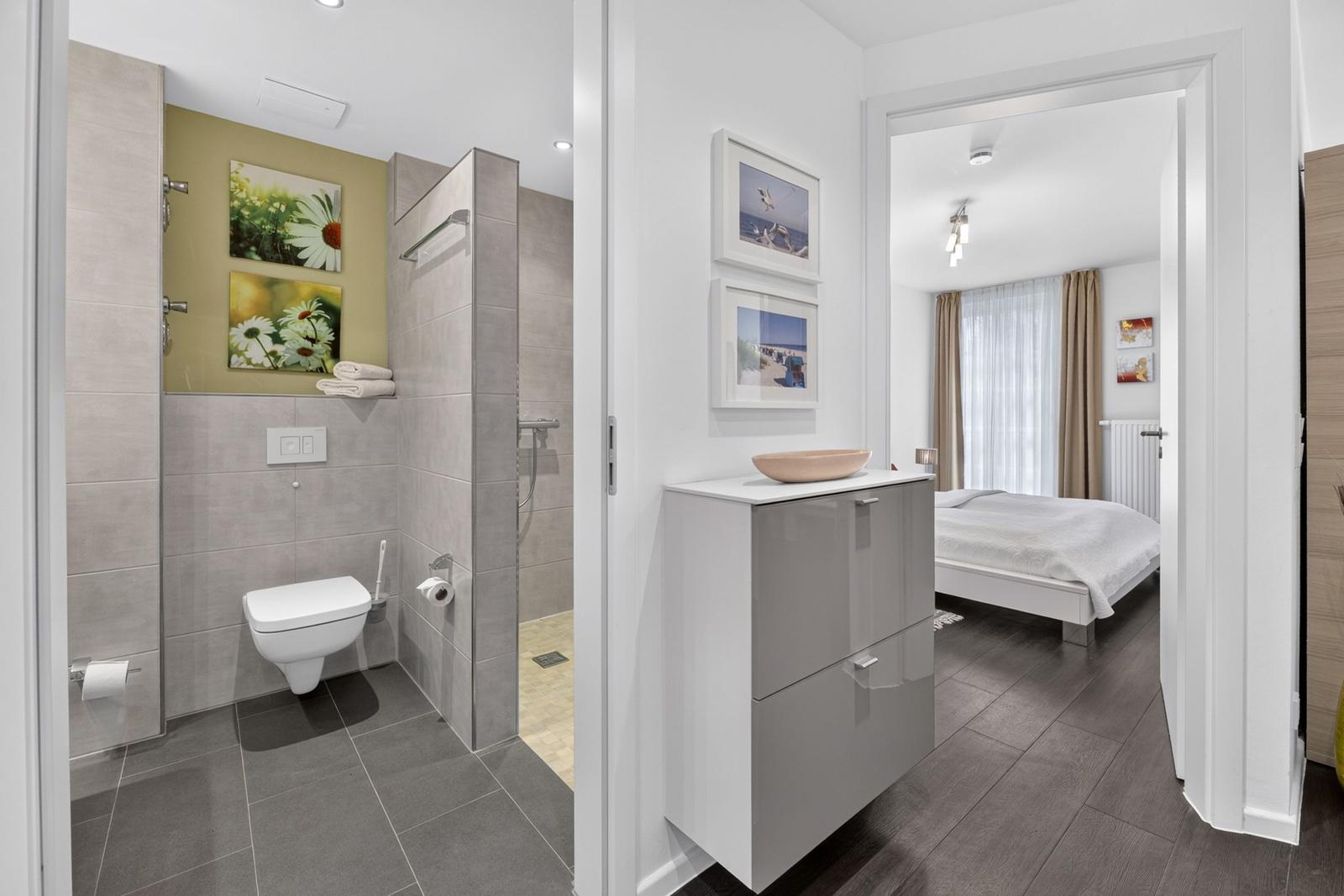 Badezimmer mit Dusche, Toilette und Waschtisch. Im Hintergrund ist ein Schlafzimmer mit Bett und Fenster.