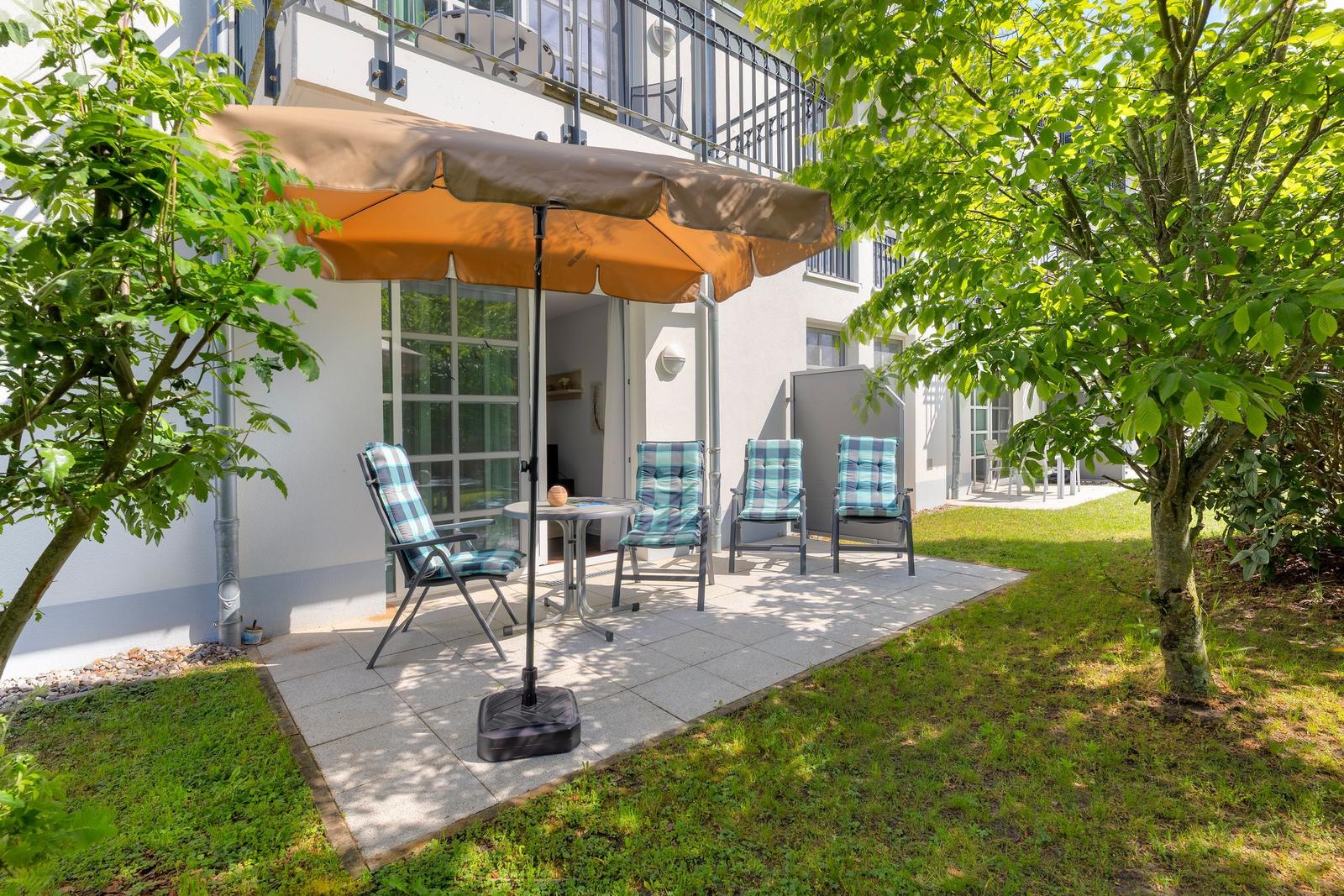 Terrasse mit Sonnenschirm, Tisch und vier Stühlen unter Bäumen.