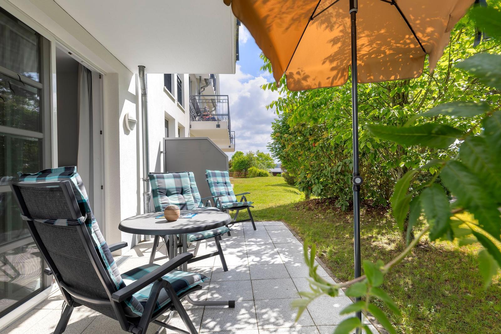 Terrasse mit Tisch, drei Stühlen und Sonnenschirm. Hintergrund: Grünflächen und Gebäude.