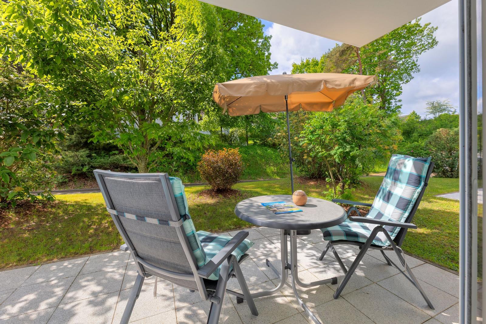 Terrasse mit Tisch, zwei Stühlen und Sonnenschirm. Grünfläche und Bäume im Hintergrund.