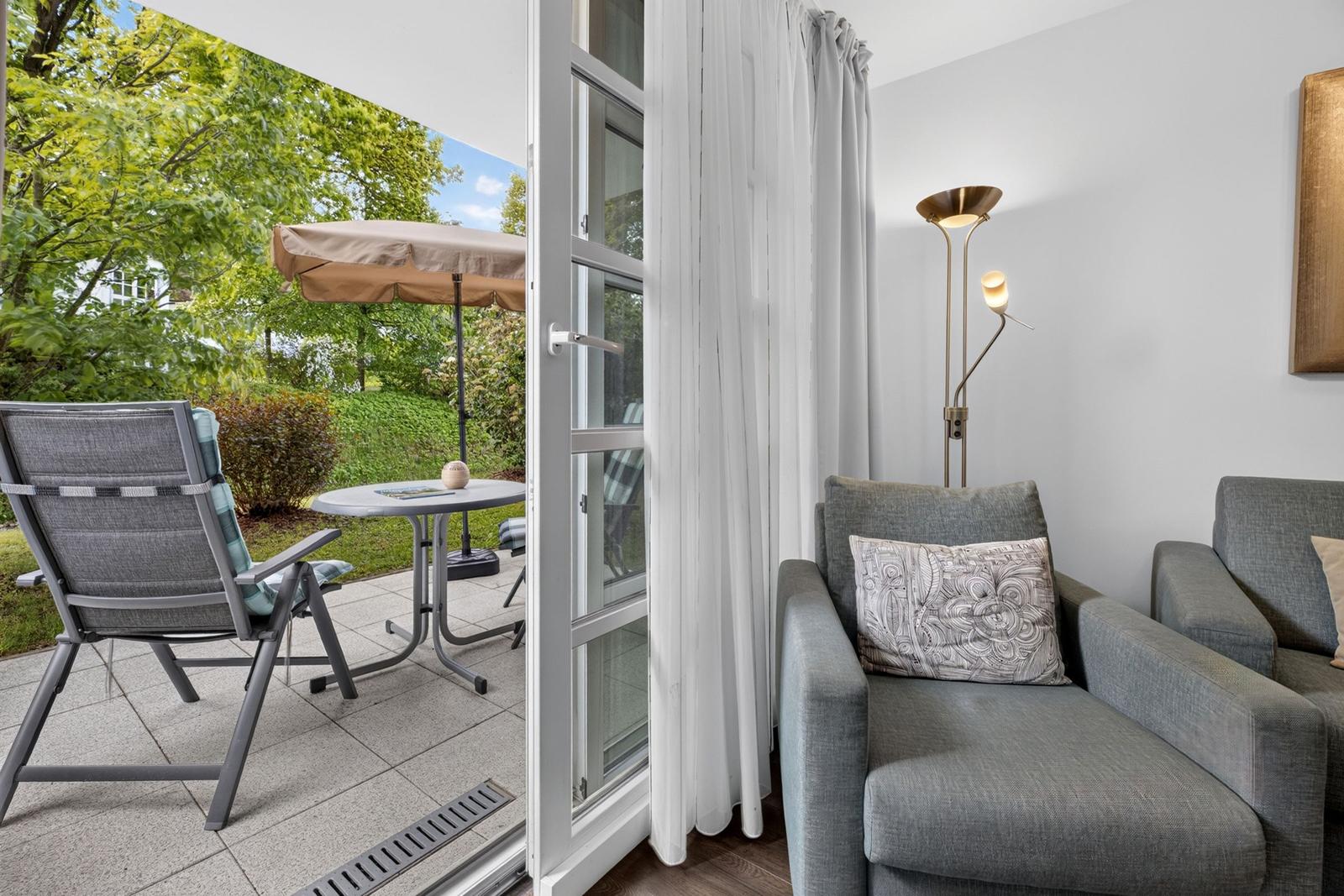 Ein Raum mit grauem Sofa und Stehlampe. Durch die Glastür ist eine Terrasse mit Stuhl und Tisch zu sehen.