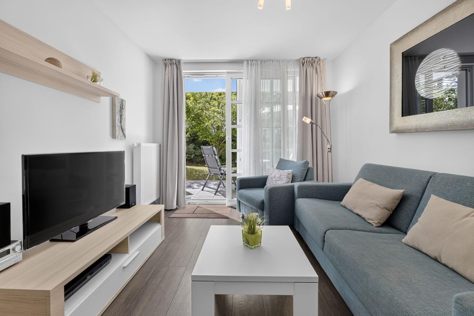 Wohnzimmer mit Sofa, Fernseher und Blick auf Balkon mit Garten.