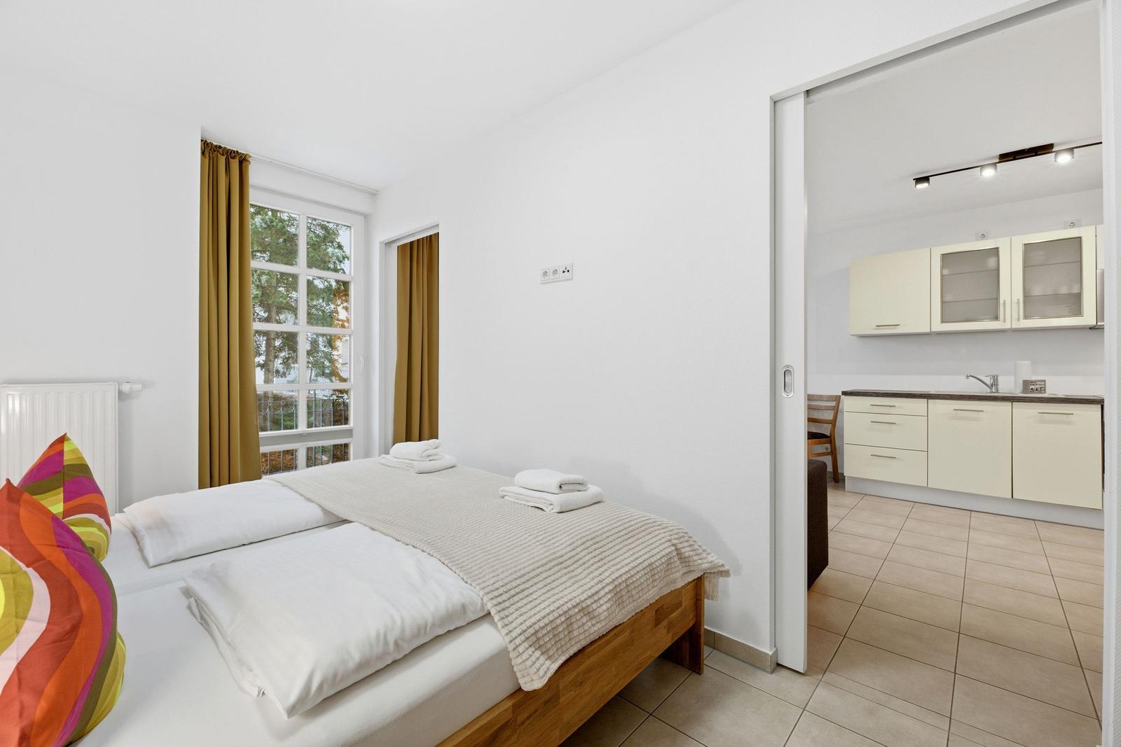 Schlafzimmer mit Bett und Fenster. Küche ist über Schiebetür sichtbar.