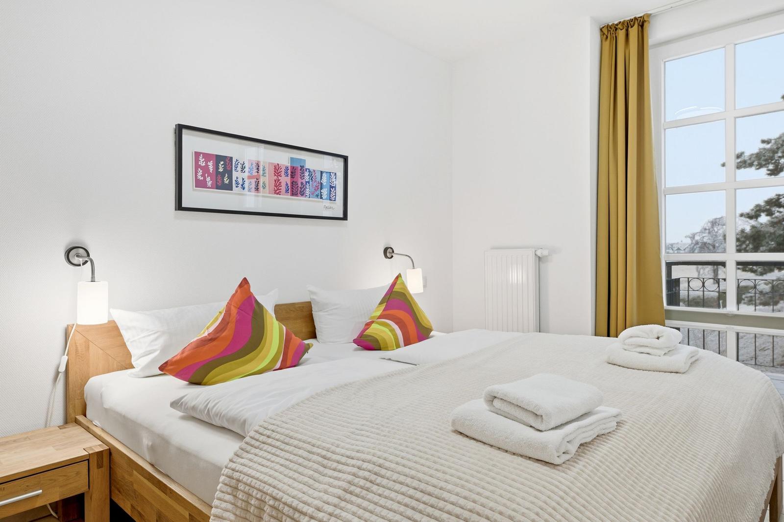 Schlafzimmer mit Doppelbett, bunten Kissen und Fenster mit Blick auf Balkon.