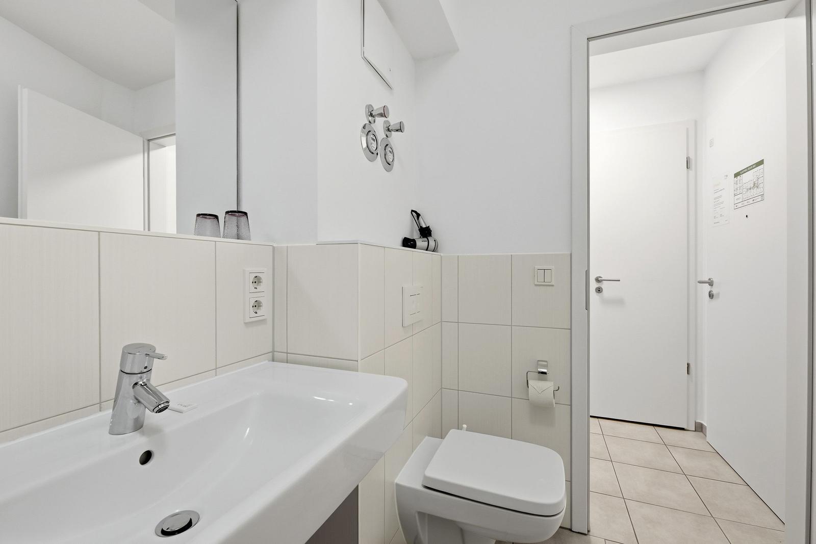 Badezimmer mit Waschbecken, Spiegel und Toilette. Fliesenwand und helle Farbgebung.