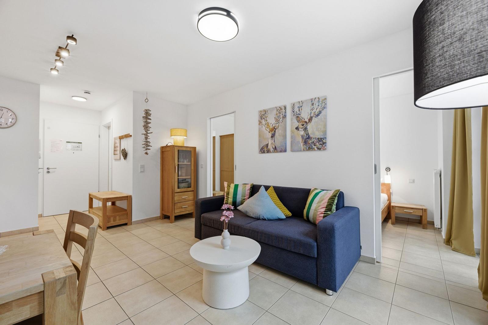 Wohnzimmer mit Sofa, Tisch und Wandbildern. Blick in Schlafzimmer mit Bett.